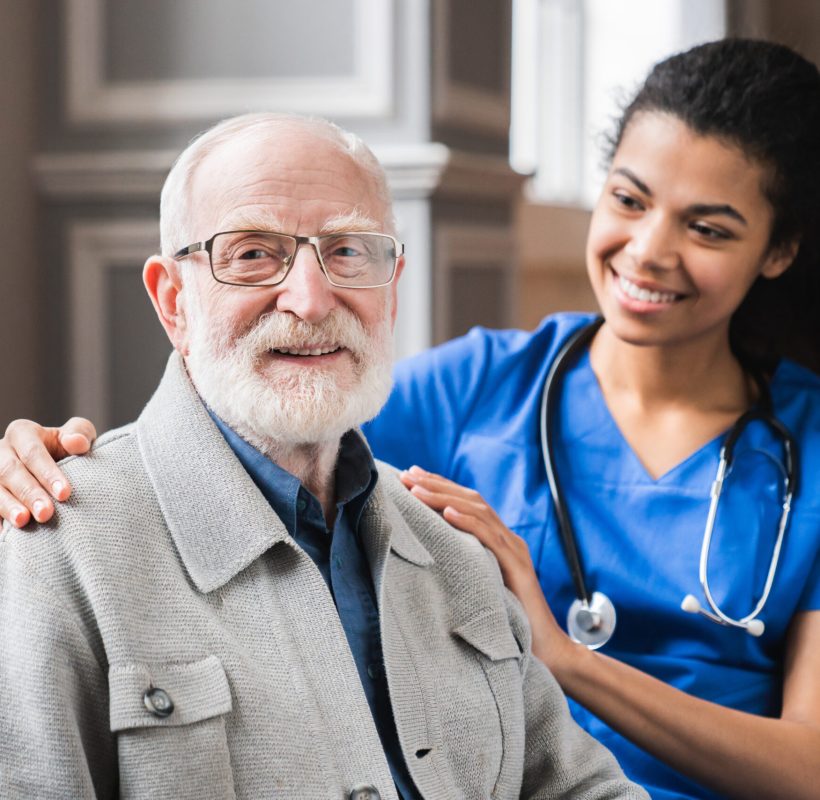 Young nurse in blue coat hugging old 80s man smiling looking at camera. Portrait of satisfied