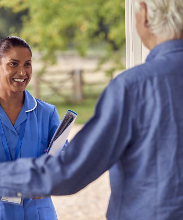 Senior Man Greeting Female Nurse Or Care Worker Making Home Visit In Uniform At Door